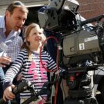 Girl operating large broadcasting camera, supervised by professional cameraman