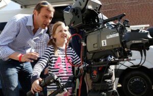 Girl operating large broadcasting camera, supervised by professional cameraman